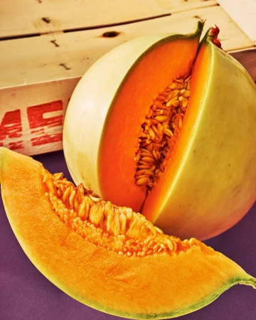 Close-up shot of a halved cantaloupe with seeds and a slice, showcasing its vibrant orange flesh against a wooden crate.
