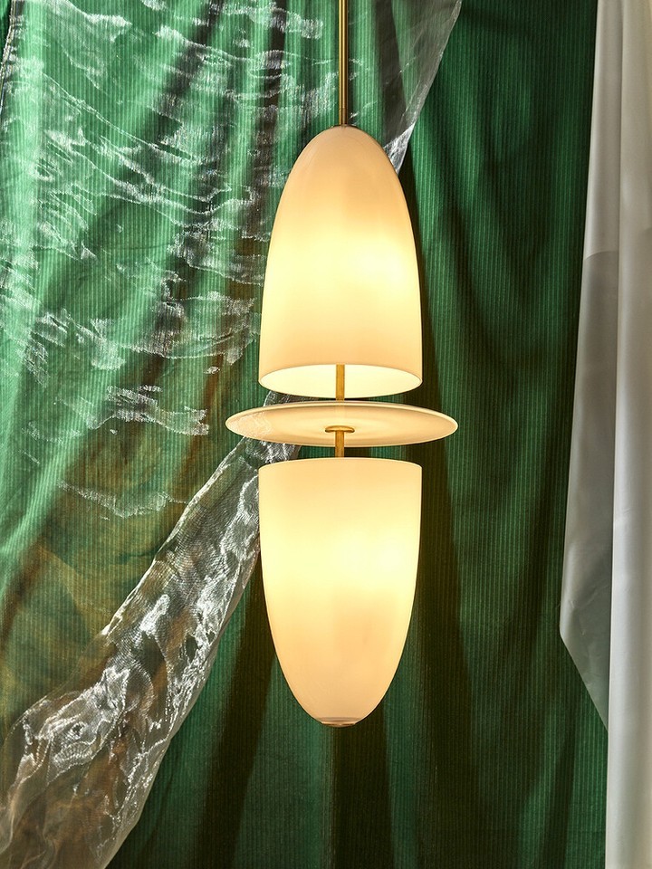 Modern pendant light fixture with a brass rod and two white glass bulb covers against a draped green fabric backdrop indoors.