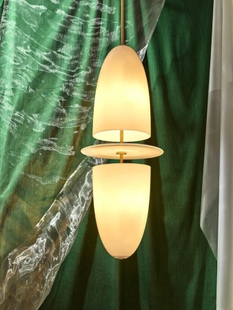 Modern pendant light fixture with a brass rod and two white glass bulb covers against a draped green fabric backdrop indoors.
