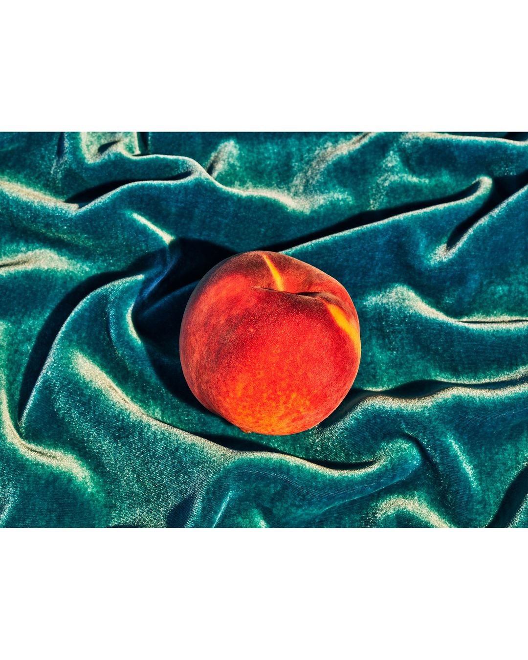 Studio shot of a fresh peach with vibrant red and yellow hues resting on a teal velvet surface with soft lighting and dramatic folds.