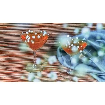 Two stemmed cocktails with light-orange liquid and white flowers are presented on a horizontal wood surface with a blue textile backdrop.