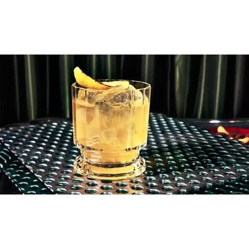 A close-up shot of a clear faceted glass filled with an orange cocktail and ice, garnished with an orange peel, sitting on a table.