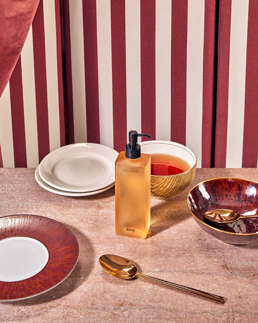 Still life shot of amber liquid soap dispenser, gold spoons, white and red plates, and bowls against a red and white striped backdrop.