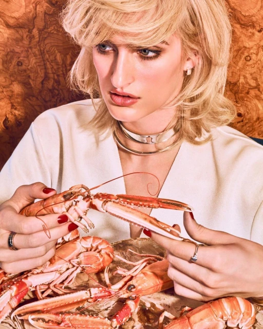 Blonde woman in fashion portrait, holds lobster on a gold platter. Wearing a white top, she poses against a textured backdrop.