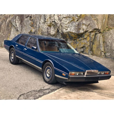 A dark blue 1980s Aston Martin Lagonda parked on an asphalt surface in front of a rocky hillside on a sunny day.