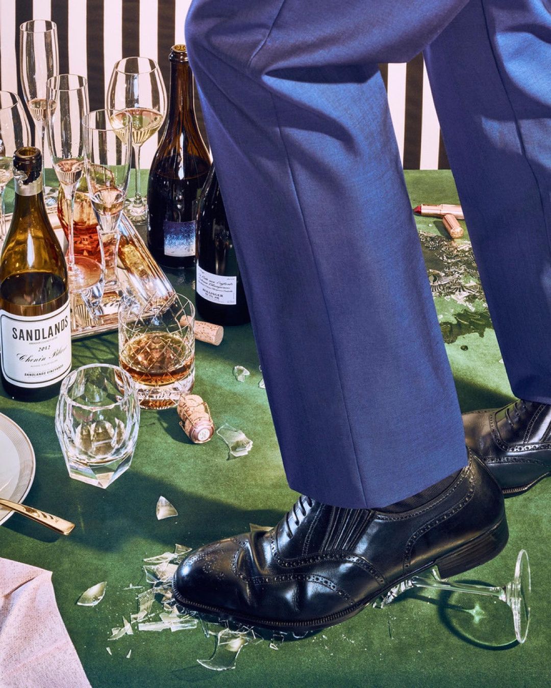 Elegant man in blue suit steps on shattered glass amidst spilled drinks and broken glassware on a green tablecloth with striped backdrop.