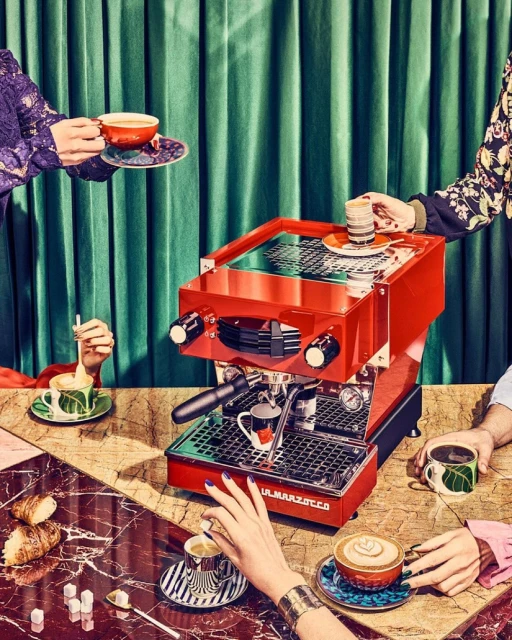 Stylish coffee presentation: red espresso machine surrounded by hands holding cups, pastries, sugar cubes on a patterned table against a green backdrop.