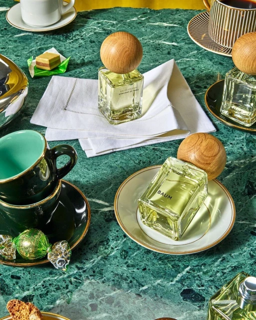 Still life shot featuring perfume bottles with wooden caps alongside cups, saucers, candies, and pastries on a green marble surface.
