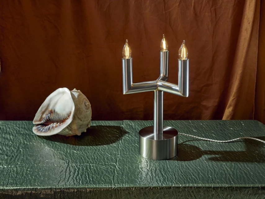Modern brushed metal table lamp with three lights and a large seashell on a green surface in front of a draped brown background.