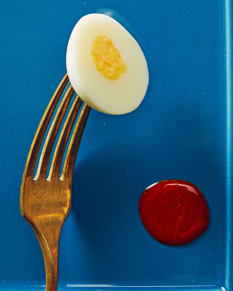 Elevated view of a halved hard-boiled egg balanced on a gold fork with a drop of red sauce on a blue plate, showcasing food photography.