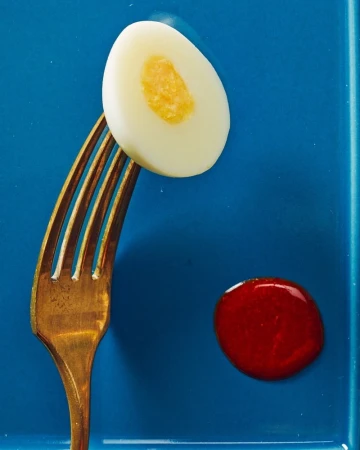 Elevated view of a halved hard-boiled egg balanced on a gold fork with a drop of red sauce on a blue plate, showcasing food photography.