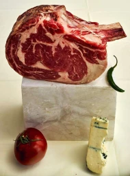 Large raw ribeye steak on bone atop a white block with a red tomato, blue cheese wedge, and green chili.