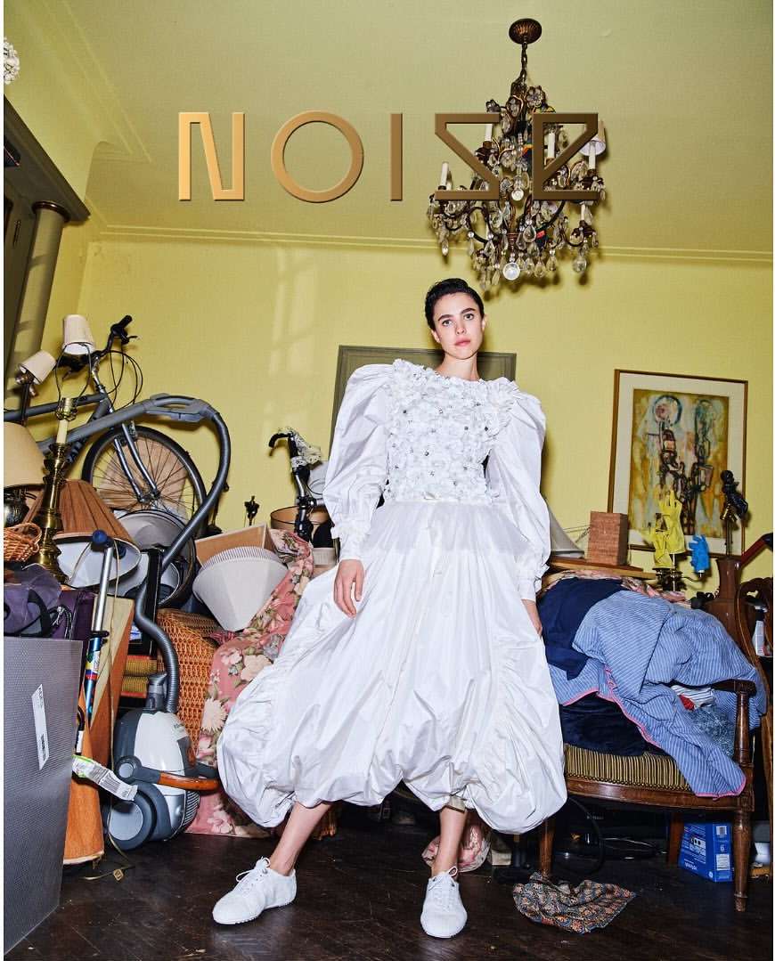 A young woman poses indoors in a voluminous white dress and sneakers, standing in a cluttered room with a chandelier and gold text above.