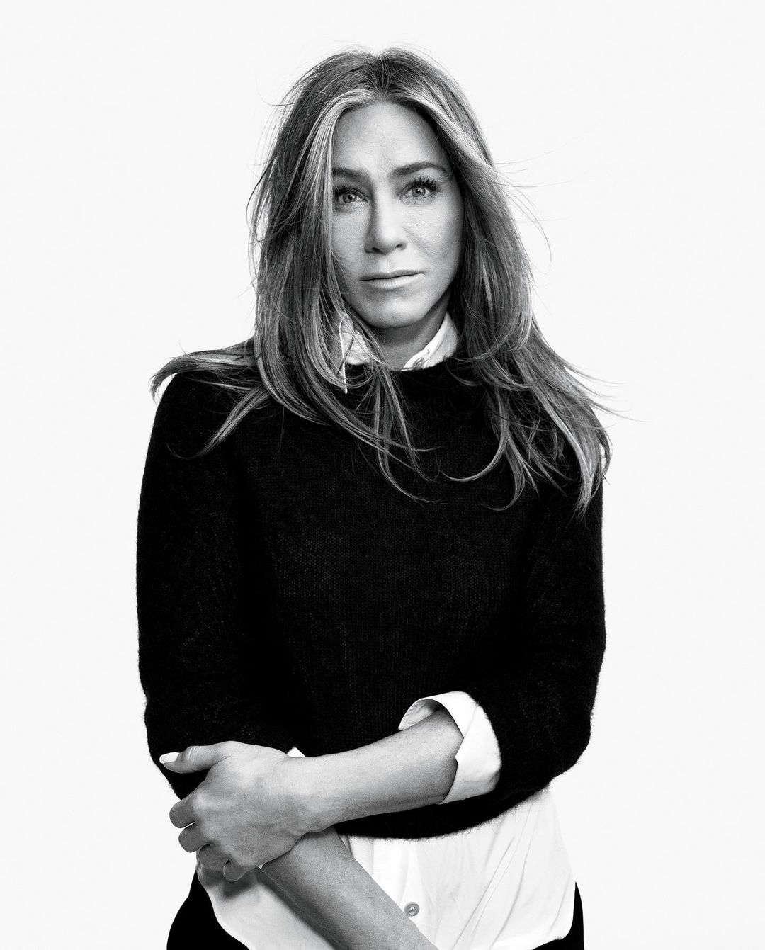 Black and white portrait of Jennifer Aniston with long brown hair and wearing a layered white collared shirt and black sweater.