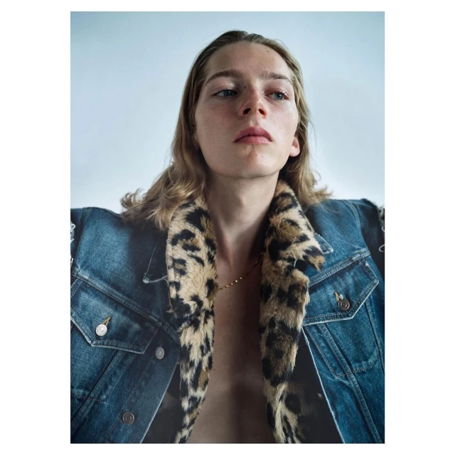 Portrait of a young adult with shoulder-length blonde hair wearing a denim jacket with a faux leopard fur collar, posing indoors against a white wall.