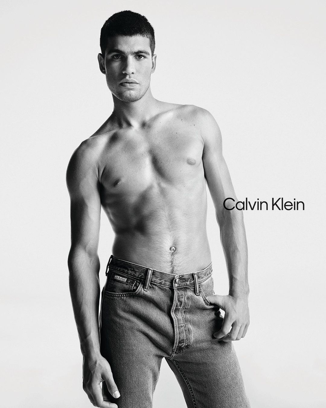 Carlos Alcaraz poses shirtless wearing blue jeans for a Calvin Klein advertisement set against a stark white backdrop in a studio.