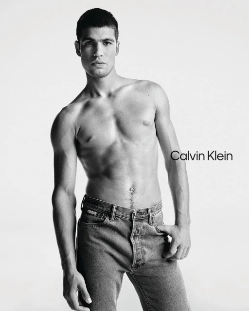 Carlos Alcaraz poses shirtless wearing blue jeans for a Calvin Klein advertisement set against a stark white backdrop in a studio.