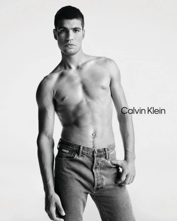 Carlos Alcaraz poses shirtless wearing blue jeans for a Calvin Klein advertisement set against a stark white backdrop in a studio.