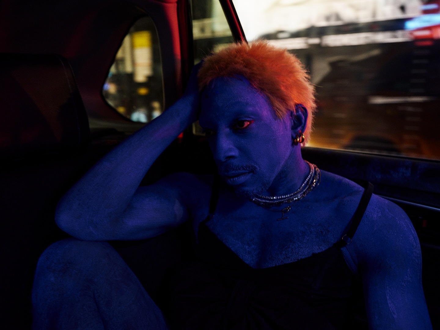 An adult with orange hair and blue body paint poses inside a car with city lights blurred in the background, illuminated by red and blue light.