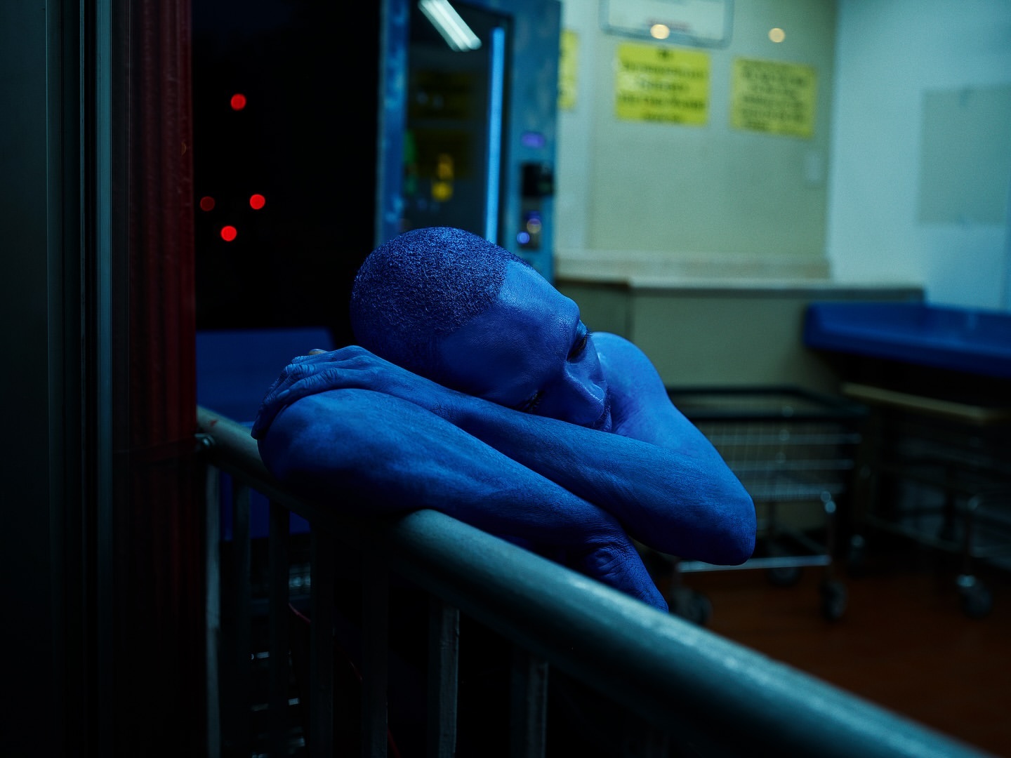 Blue-skinned adult rests head on railing indoors, bathed in cool, artificial lighting. Sparse environment suggests public space, evoking pensive mood.