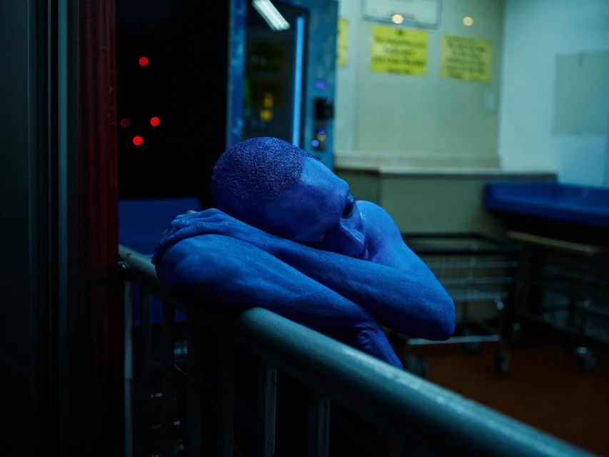 Blue-skinned adult rests head on railing indoors, bathed in cool, artificial lighting. Sparse environment suggests public space, evoking pensive mood.