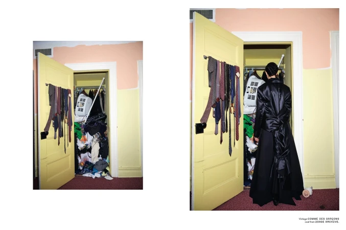 Woman stands before an open closet in a room with yellow and pink walls, filled with assorted clothes, ties hung on the door.