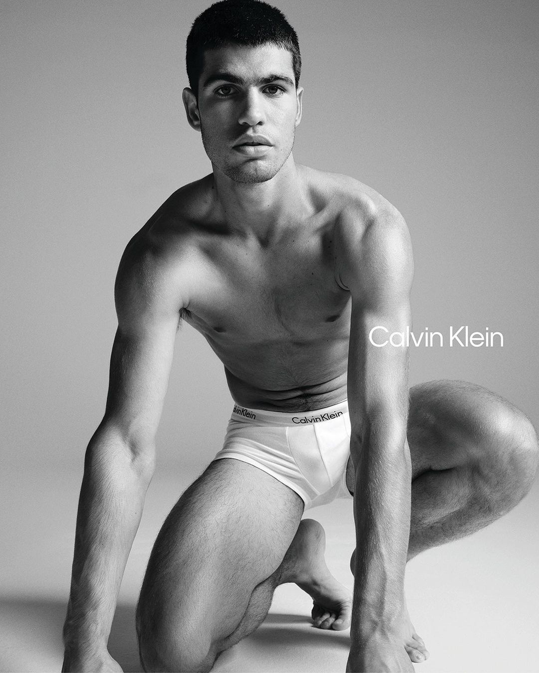 Black and white portrait of shirtless man kneeling in Calvin Klein underwear against grey background, showcasing fashion and fitness.