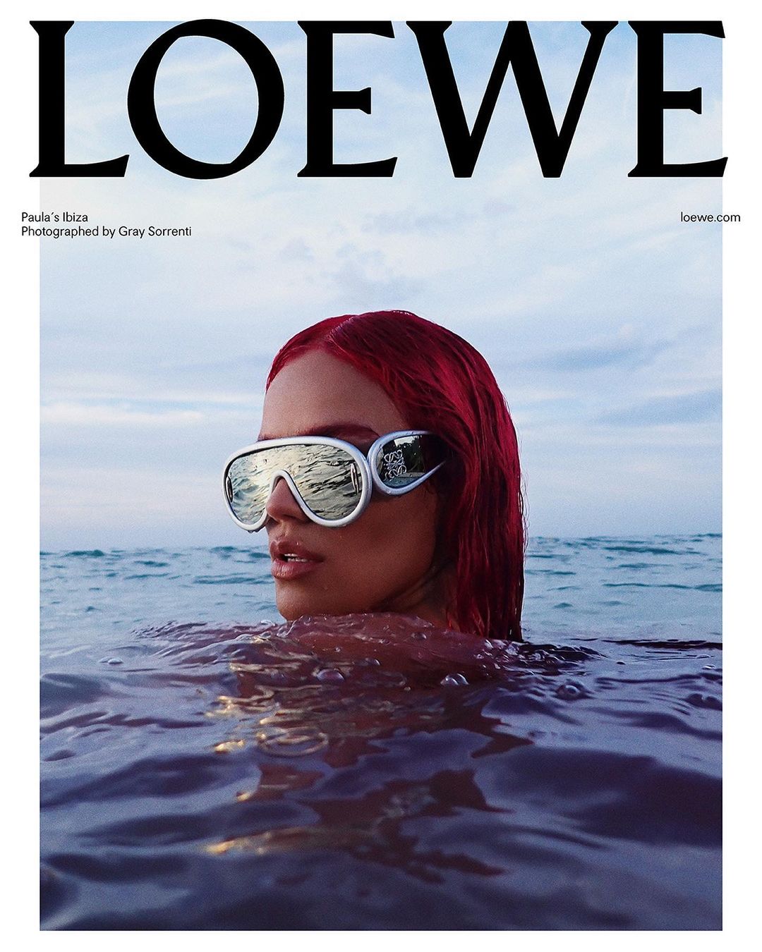 Close-up of a woman in water wearing futuristic sunglasses, red hair, against a background of open water and a light blue sky.  