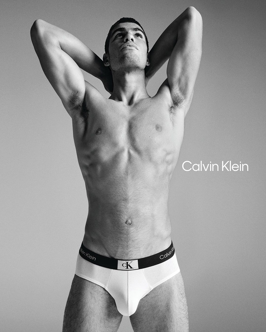 Male fashion model in white Calvin Klein underwear poses shirtless against a grey studio background with logo text.