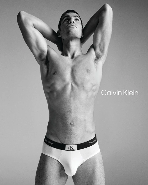 Male fashion model in white Calvin Klein underwear poses shirtless against a grey studio background with logo text.