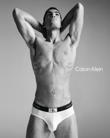 Male fashion model in white Calvin Klein underwear poses shirtless against a grey studio background with logo text.