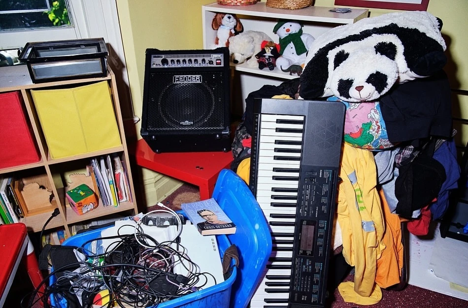 Indoor cluttered room with a keyboard, speaker, storage shelves, and disorganized household items like clothes and toys.