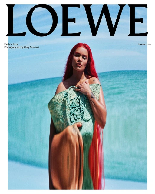 A woman with red hair poses near ocean wearing a green woven Loewe bag. Fashion editorial shot with blue sky and sea backdrop.