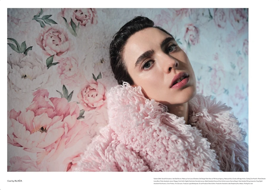 Portrait of a woman with dark hair wearing a pink faux fur coat against a floral background, looking up and to the right.