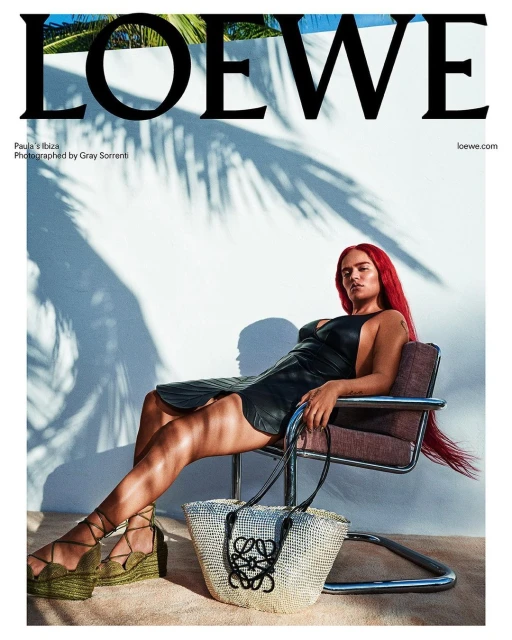 Editorial fashion shot of a woman with red hair wearing a black dress, sitting on a chair with a tote bag at her feet.