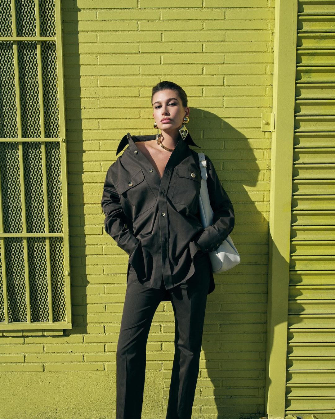 Adult female poses in fashionable black shirt and pants, accessorized with large earrings and a shoulder bag, against a yellow brick wall.