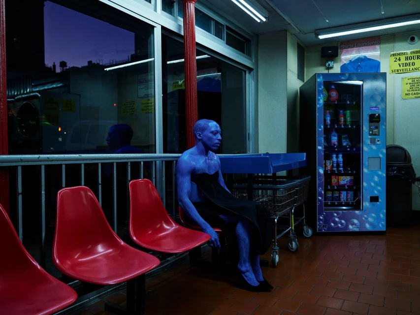 Blue-skinned adult sits pensively on a red plastic chair in a brightly lit laundromat, with a vending machine in the background.