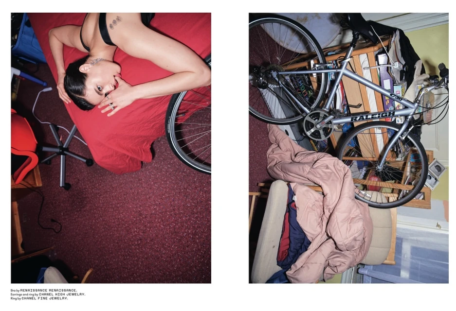 Woman lying on a red bed next to a bicycle in an indoor setting with a patterned carpet. Soft lighting emphasizes the scene.