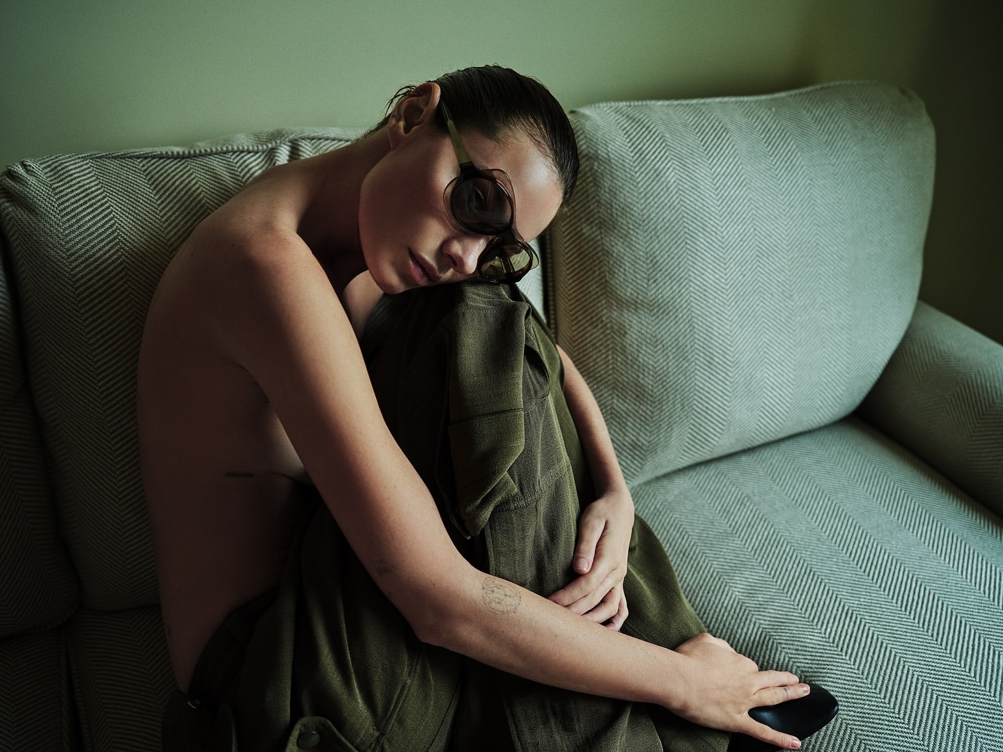 Adult female posing on a light herringbone couch indoors wearing sunglasses and an olive green pantsuit in a portrait format image.
