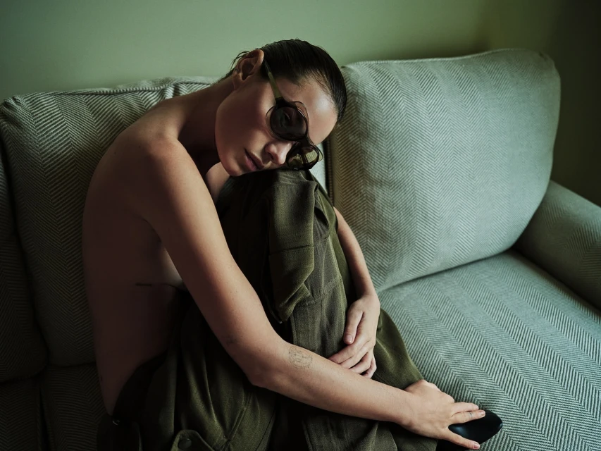 Adult female posing on a light herringbone couch indoors wearing sunglasses and an olive green pantsuit in a portrait format image.