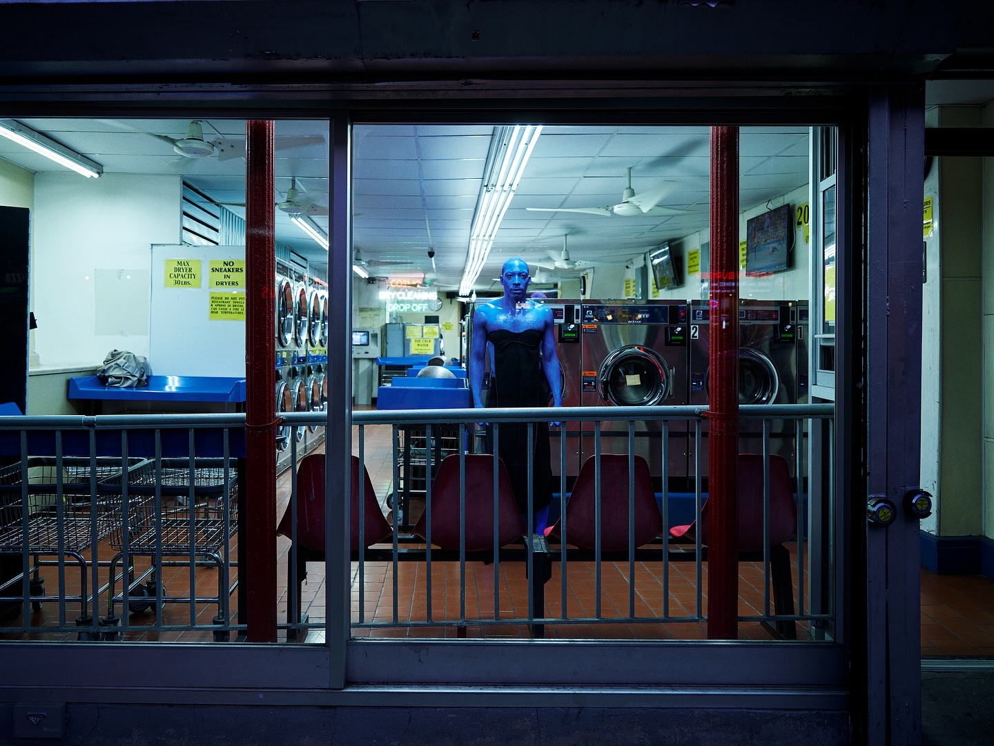 An adult human with bright blue skin and black attire stands in a laundromat with rows of washing machines and metal seating.