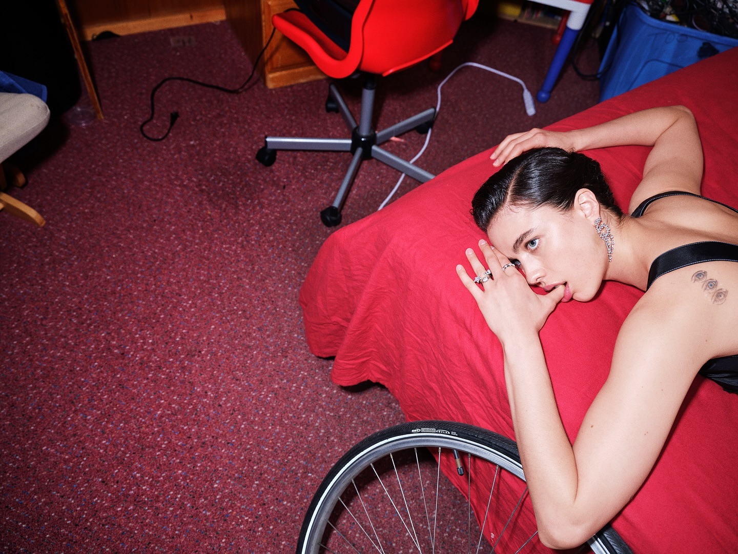 Fashion shot of a woman in lingerie lying on a red bed with a bicycle wheel in the foreground. Red chair and carpet visible.