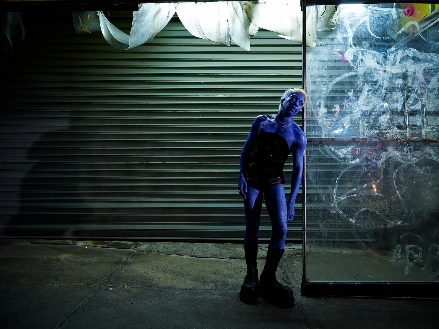 Blue-skinned person in corset and boots strikes a pose against a grungy urban backdrop of closed metal shutter and graffiti-covered glass.
