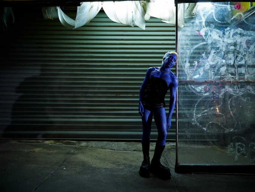 Blue-skinned person in corset and boots strikes a pose against a grungy urban backdrop of closed metal shutter and graffiti-covered glass.