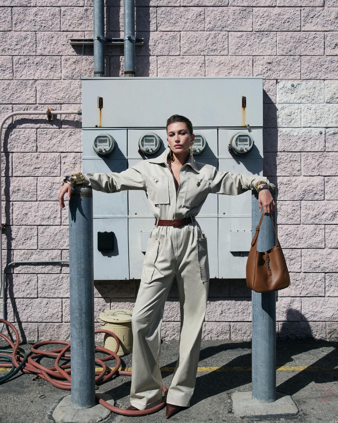 A woman in a tan jumpsuit leans against poles outside a building with electrical boxes, accessorized with a brown belt and purse.