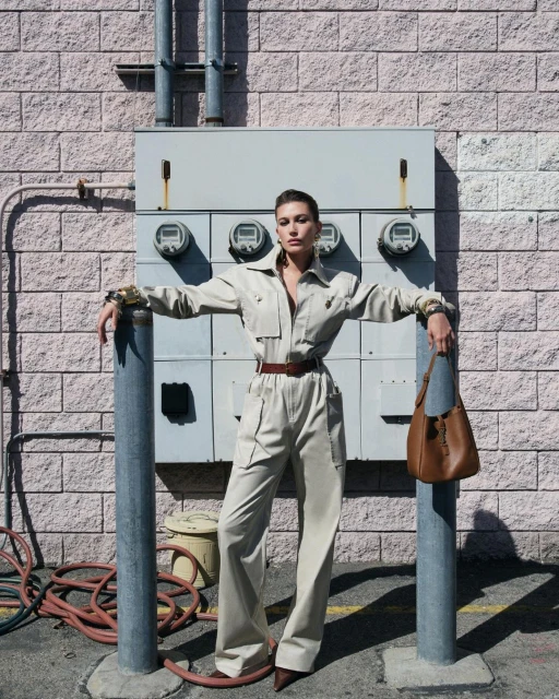 A woman in a tan jumpsuit leans against poles outside a building with electrical boxes, accessorized with a brown belt and purse.