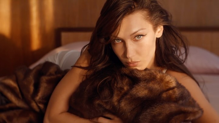Attractive woman in a low-angle portrait lying on bed with a fur blanket, gazing directly at the camera with a serious expression indoors.