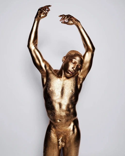 Gold-painted adult male portrait with hands raised. Bronze skin, eyes closed, in a photo studio against a neutral light background.