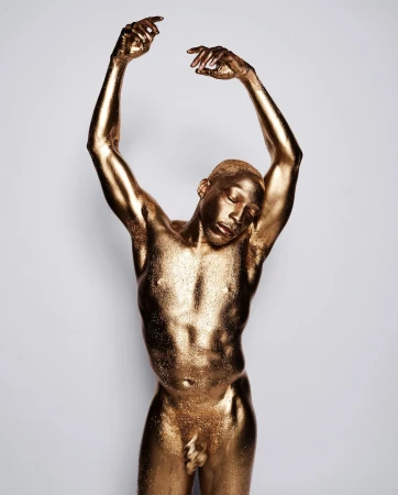 Gold-painted adult male portrait with hands raised. Bronze skin, eyes closed, in a photo studio against a neutral light background.