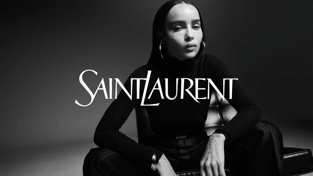 Black and white studio portrait of an adult female model wearing a black turtleneck and silver hoop earrings, with 'Saint Laurent' logo overlay.